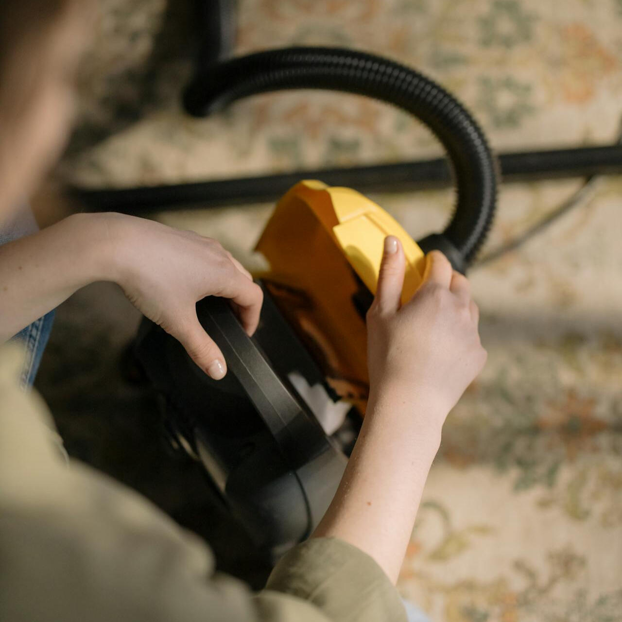 Osoba otwiera żółty odkurzacz na dywanie.